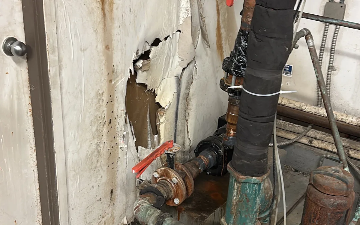 Torn, water-stained wallboard beside corroded piping and valves in a mechanical room
